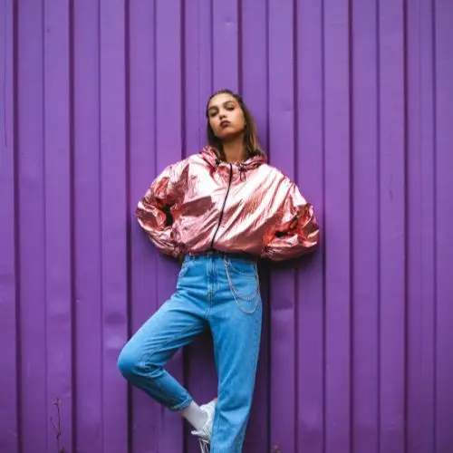  A woman in a metallic jacket and jeans leans casually against a vibrant purple wall, showcasing a modern urban style.