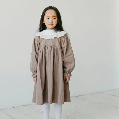 A little girl dressed in a brown dress and white tights poses happily, showcasing her playful spirit and youthful charm.<br />
