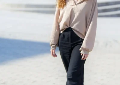 A woman wearing a beige sweater and black pants stands confidently, showcasing a casual yet stylish outfit.