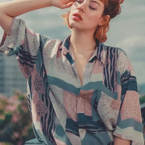 A woman in a vibrant shirt poses confidently on a balcony, showcasing her style against a scenic backdrop.